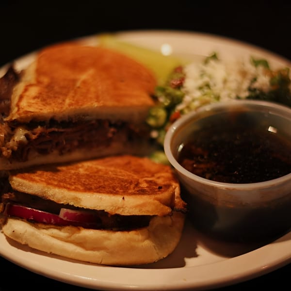 Grilled sandwich with meat and cheese, a side salad, and a cup of dark sauce on a white plate.