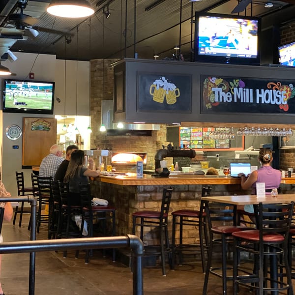 Diners at The Mill House bar with a wood-fired pizza oven and TVs showing sports.