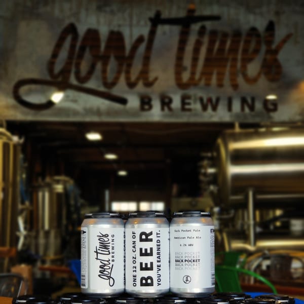 Three cans of "Good Times Brewing" beer in front of brewing equipment and a blurred sign.