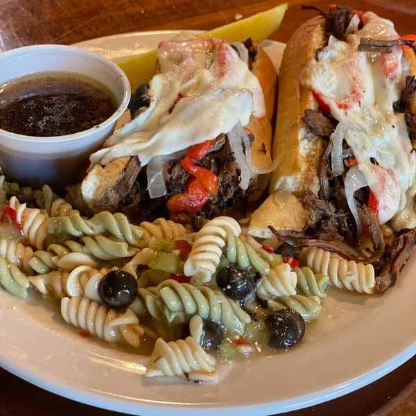 Two cheesesteak sandwiches with a side of pasta salad and a cup of au jus.