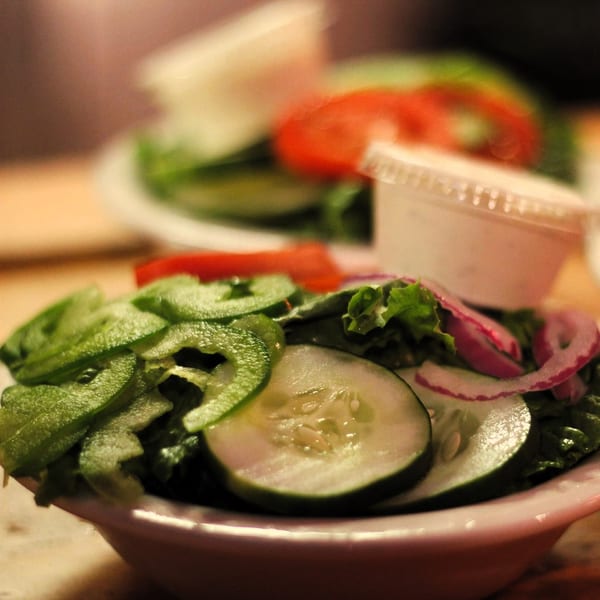 Two side salads with lettuce, cucumbers, red onions, green peppers, and tomatoes, with dressing cups.